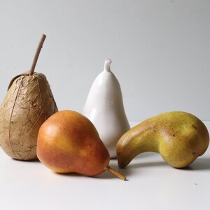 May include: Still life composition featuring four pear-shaped objects. A pear wrapped in textured brown material, a smooth orange pear, a white ceramic pear, and a green and yellow pear with a curved neck.