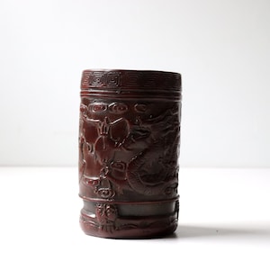 May include: A dark red cylindrical vase with intricate carvings of dragons and other mythical creatures. The vase has a textured surface and a slightly flared lip.