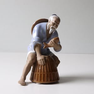 May include: A ceramic figurine of a man in a blue robe holding a fish. He is sitting on a brown woven basket.