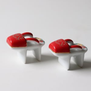 May include: Two ceramic Japanese geta, miniature versions, are displayed. The geta feature a white base with three legs, a red top with a gold design, and a red strap. Floral patterns decorate the sides. The geta are likely decorative.