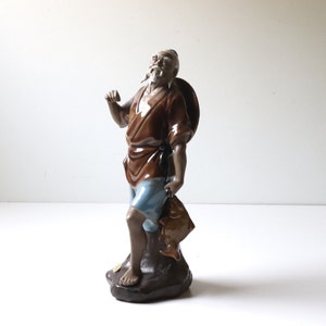 May include: A ceramic figurine of a man holding a fish. He is wearing a brown robe and blue pants. He is standing on a brown base.