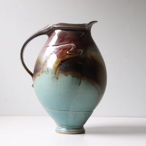 May include: A handcrafted ceramic pitcher with a large handle and a spout. The pitcher features a two-tone design, with a turquoise base and a brown and maroon upper section. The surface has a glossy finish, and the pitcher is likely used for serving beverages.