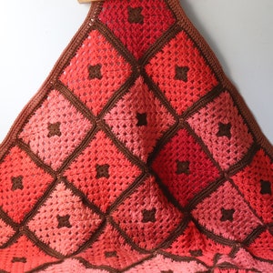 May include: A crocheted blanket with a gradient of red, pink, and brown squares. The blanket is made with a simple stitch pattern and has a brown border.