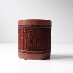 May include: A cylindrical wooden container with a reddish-brown finish. The container is decorated with a repeating pattern of black lines and Chinese characters.