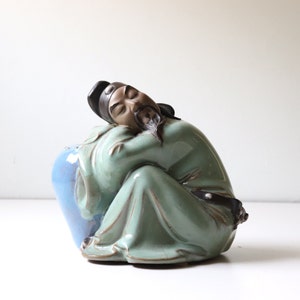 May include: A ceramic figurine of a man in a green robe sitting with his arms crossed and a blue and green vase at his feet.