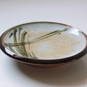 May include: A small, white ceramic dish with a brown rim and green and brown brushstrokes. The dish has a slightly textured surface.