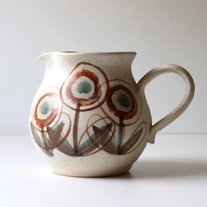 May include: A cream-colored ceramic pitcher with a speckled finish, featuring a hand-painted design of three stylized flowers in shades of rust-orange, brown, and muted green. The pitcher has a rounded body, a curved handle, and a small pouring spout.