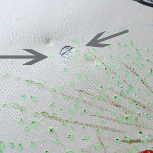 May include: A close-up of a white canvas with a hole in the center. The canvas is covered in small green and gold paint splatters. Two arrows point to the hole.