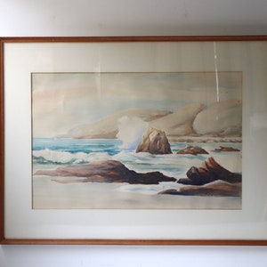 May include: A framed watercolor painting depicting a seascape with crashing waves, rocky cliffs, and a blue sky. The painting is in a light brown wooden frame.