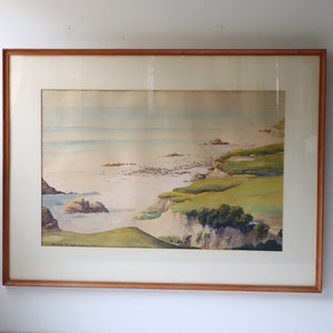 May include: Framed watercolor painting depicting a coastal scene with a rocky shoreline, green grassy hills, and a calm blue ocean in the background.
