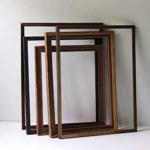 May include: A collection of four rectangular picture frames stacked behind one another. The frames vary in color, including dark brown, black, and light brown wood tones, and feature simple, clean edges without glass or backing inserts.