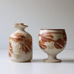 May include: A ceramic cruet and a matching stemmed goblet, both featuring a speckled off-white glaze decorated with reddish-brown leaf imprints. The cruet has a narrow neck and a small spout, while the goblet has a rounded bowl and a pedestal base.