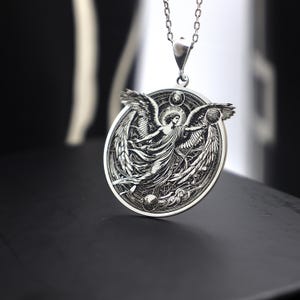 May include: A silver pendant necklace featuring an intricate design of an angel with outstretched wings. The angel is surrounded by a circular frame with celestial symbols.