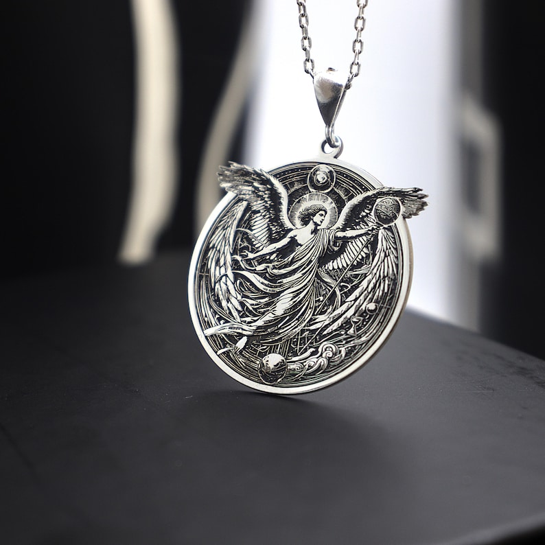 May include: A silver pendant necklace featuring an intricate design of an angel with outstretched wings. The angel is surrounded by a circular frame with celestial symbols.