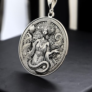 Può includere: Ciondolo in argento con un design intricato di una donna che tiene una brocca, circondata da motivi a spirale e un serpente. Il ciondolo è circolare e ha un bordo dettagliato e ornato.