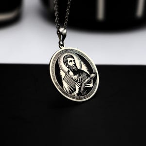 Op de afbeelding: Zilveren hangerketting met een ovale hanger met een gravure van een religieuze figuur met een boek. De hanger hangt aan een fijne zilveren ketting. De achtergrond is zwart en wit.