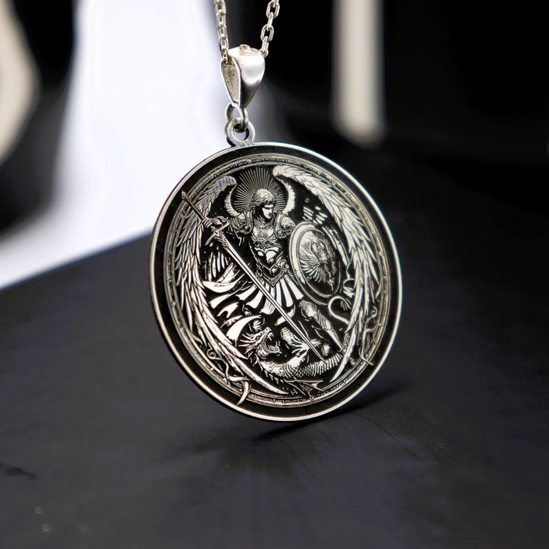 May include: Silver pendant necklace featuring a detailed engraving of an angel with wings spread wide, holding a sword and shield, standing over a dragon. The pendant is circular and has a black background.