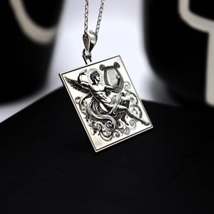 Puede incluir: Collar con colgante de plata con un dije de forma cuadrada grabado con una imagen de un ángel tocando la lira.