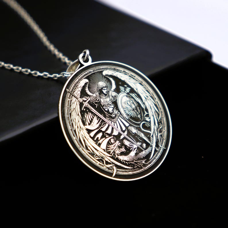 May include: Silver pendant necklace featuring a detailed engraving of an angel with wings, a sword, and a shield. The angel is standing over a dragon.