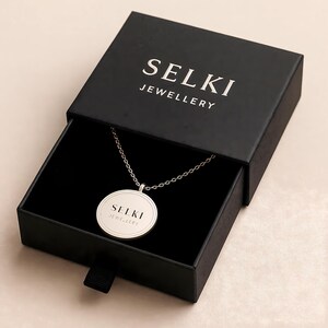A necklace with a round pendant is displayed in a black jewelry box with the brand name "SELKI JEWELLERY" printed on it.