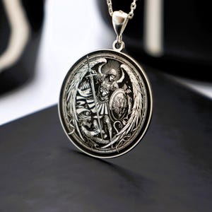 May include: Silver pendant necklace featuring a detailed engraving of an angel with wings, holding a sword and shield, fighting a dragon. The pendant is round and has a polished finish.