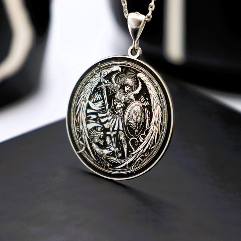 May include: Silver pendant necklace featuring a detailed engraving of an angel with wings, holding a sword and shield, fighting a dragon. The pendant is round and has a polished finish.