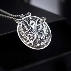 May include: Silver pendant necklace featuring an angel with wings outstretched, holding a sphere. The angel is surrounded by a circular design with celestial bodies.