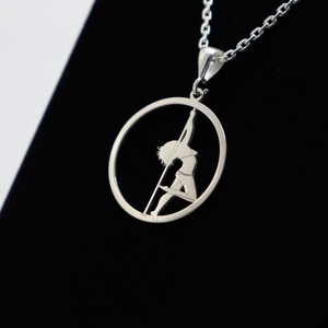 Può includere: Collana con ciondolo in argento con un design a taglio di una persona in un cerchio. La persona è in silhouette e sembra eseguire una danza del palo.