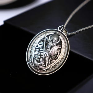 May include: A silver pendant necklace featuring an engraved image of an angel with wings, holding a sword and shield. The angel is depicted in a battle scene, with a dragon at its feet.