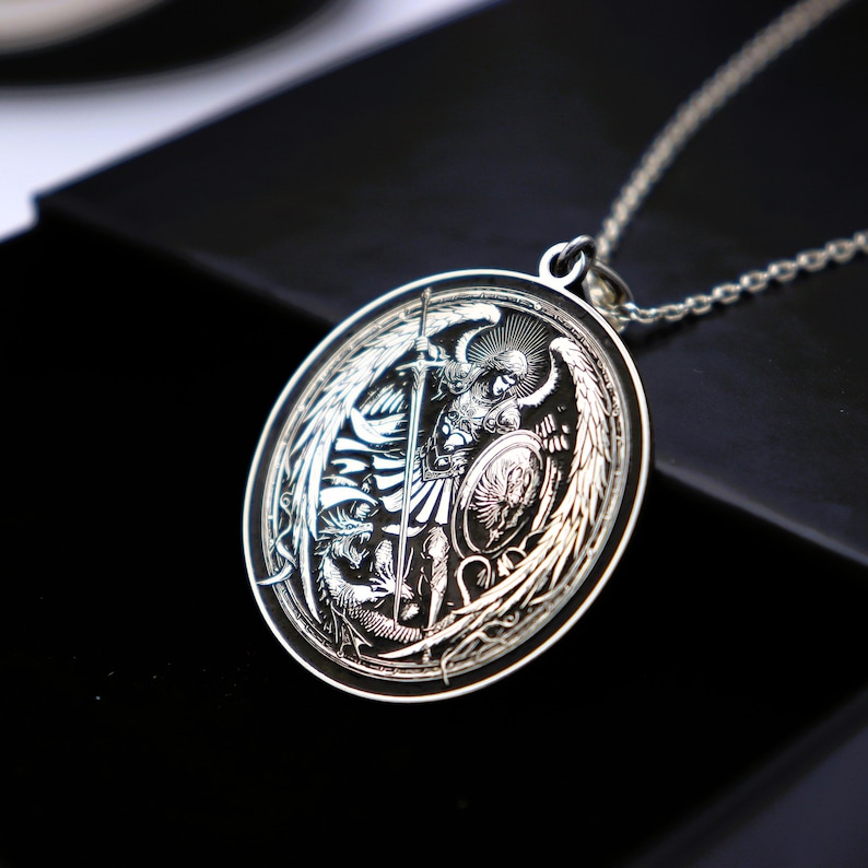 May include: A silver pendant necklace featuring an engraved image of an angel with wings, holding a sword and shield. The angel is depicted in a battle scene, with a dragon at its feet.