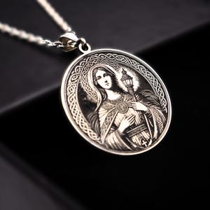 A silver pendant featuring an engraved image of a woman, possibly a religious figure, with intricate patterns surrounding her.