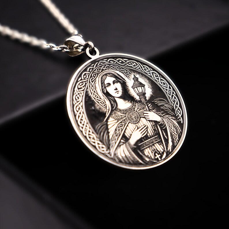 A silver pendant featuring an engraved image of a woman, possibly a religious figure, with intricate patterns surrounding her.