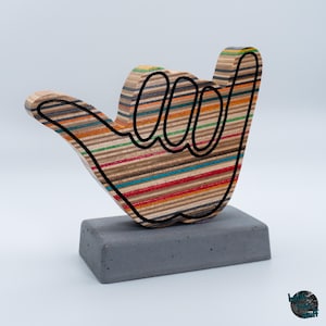 Könnte beinhalten: Eine Skulptur in Form einer Shaka-Handgeste, gefertigt aus Schichten von mehrfarbigem, recyceltem Skateboard-Holz mit einer markanten schwarzen Umrandung. Die Handfigur ist auf einem soliden, rechteckigen grauen Betonsockel montiert.