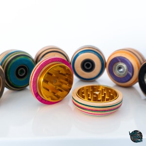 Puede incluir: Una colección de molinillos de hierbas hechos de madera laminada multicolor con interiores de metal dorado que presentan dientes afilados. Los molinillos son circulares, algunos muestran rodamientos de bolas expuestos y están agrupados.
