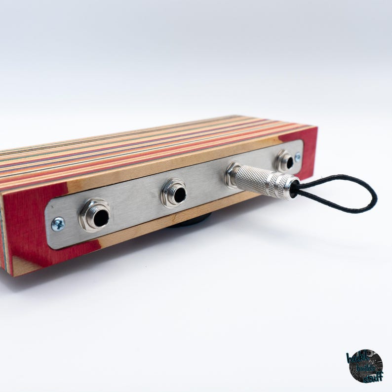 May include: A rectangular audio device made from layered wood with red, brown, and tan tones. It features a metal plate with three input jacks and a silver-colored plug with a black loop. The device is set against a white background.