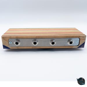 May include: A rectangular wooden audio device with a striped top in shades of red, yellow, and brown. It features a silver metal plate with four input jacks. The sides are made of wood and have a blue accent.