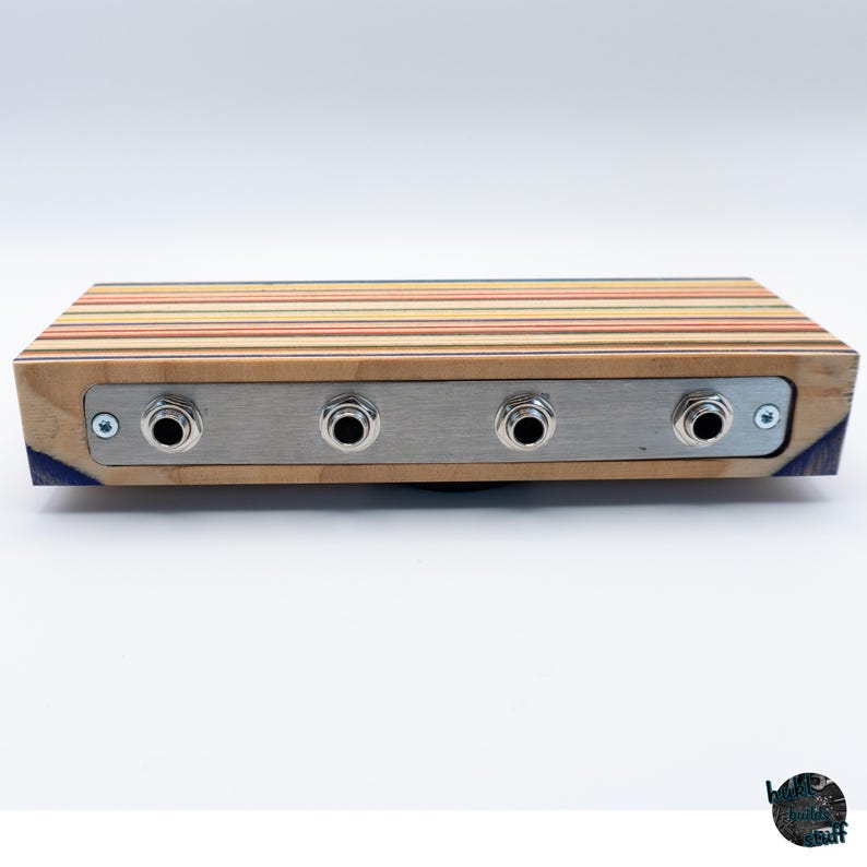 May include: A rectangular wooden audio device with a striped top in shades of red, yellow, and brown. It features a silver metal plate with four input jacks. The sides are made of wood and have a blue accent.