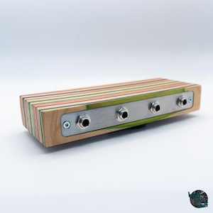 May include: A rectangular audio device with a wooden body made from layered skateboard decks. It features a brushed metal faceplate with four input jacks. The wood has stripes of various colors, including green, red, and blue.