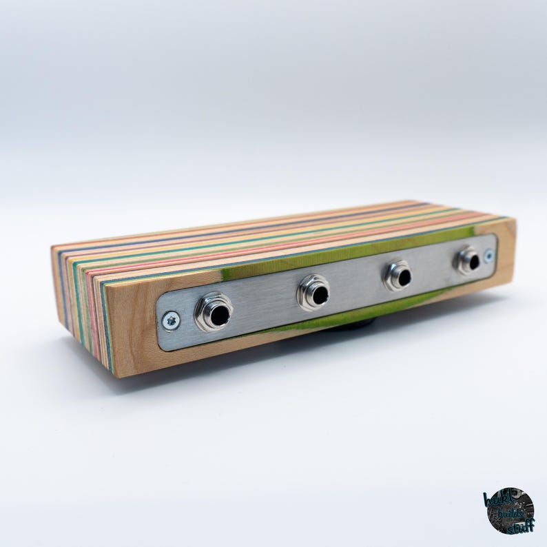 May include: A rectangular audio device with a wooden body made from layered skateboard decks. It features a brushed metal faceplate with four input jacks. The wood has stripes of various colors, including green, red, and blue.