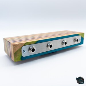May include: A rectangular wooden audio device with a metal faceplate. The wood has layers of blue, green, and natural tones. The metal plate has three input jacks. The device is designed for audio equipment.