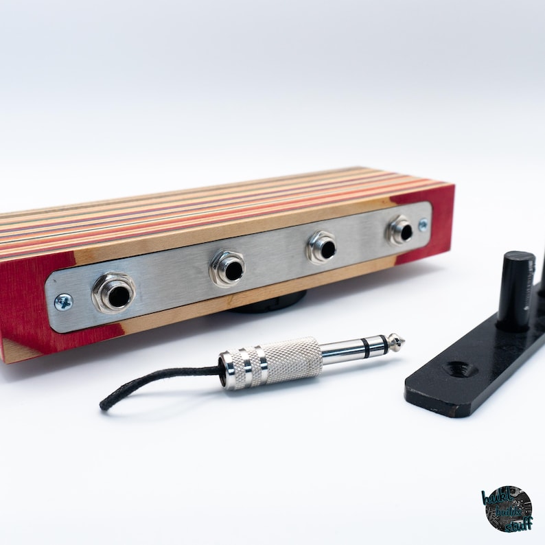 May include: A wooden audio device with a metal faceplate and three input jacks. The device is made of layered wood in shades of red, orange, and natural wood tones. A silver audio jack and a black stand are also present.