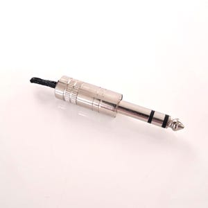 May include: A silver-colored 1/4 inch (6.35 mm) audio connector with a black cable attached. The connector has a cylindrical body with ridges and a tip with two black bands.