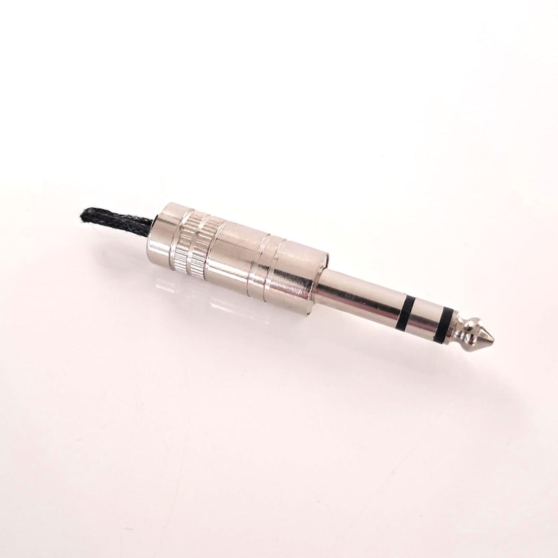 May include: A silver-colored 1/4 inch (6.35 mm) audio connector with a black cable attached. The connector has a cylindrical body with ridges and a tip with two black bands.