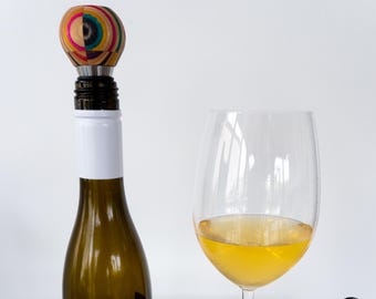 May include: A colorful wooden bottle stopper with a silver top, on top of a bottle of white wine, next to a glass of white wine.