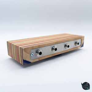 May include: A rectangular wooden audio device with a multi-colored striped design. It features a silver metal plate with three input jacks. The device is likely for audio equipment or musical instruments.