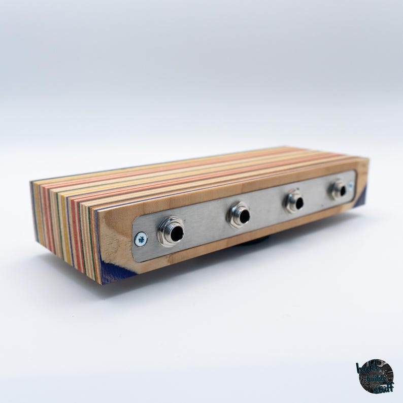 May include: A rectangular wooden audio device with a multi-colored striped design. It features a silver metal plate with three input jacks. The device is likely for audio equipment or musical instruments.