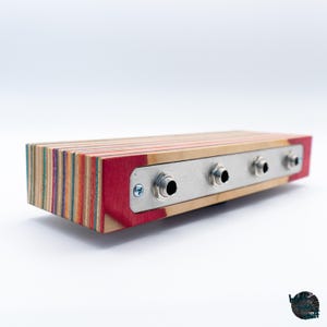 May include: A rectangular audio device with a multi-colored wood grain body and a red end. It features a silver metal plate with four input jacks. The device is designed for audio equipment.