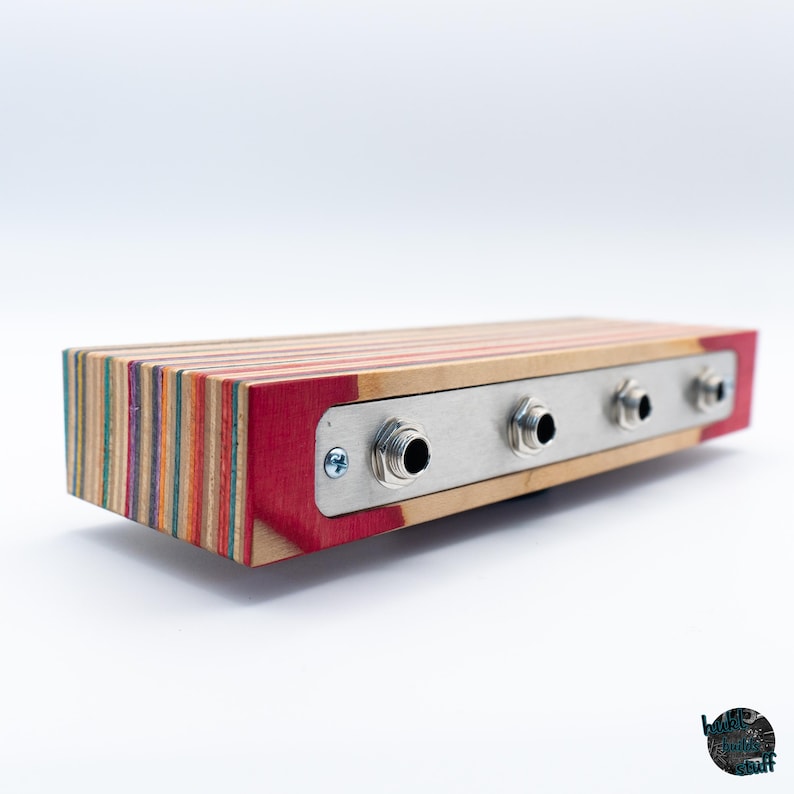 May include: A rectangular audio device with a multi-colored wood grain body and a red end. It features a silver metal plate with four input jacks. The device is designed for audio equipment.