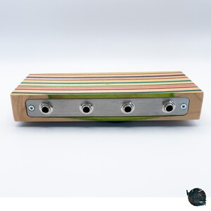 May include: A rectangular wooden audio device with a multi-colored striped top and a silver metal panel. The panel has four input jacks. The sides of the device are light-colored wood.