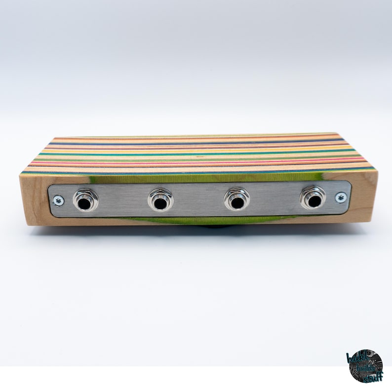 May include: A rectangular wooden audio device with a multi-colored striped top and a silver metal panel. The panel has four input jacks. The sides of the device are light-colored wood.